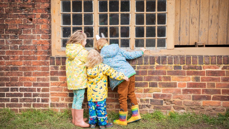 Three young children wearing colourful raincoats and wellies peer through the window of one of the stableyards buildings as part of the Easter trail at Calke Abbey.
